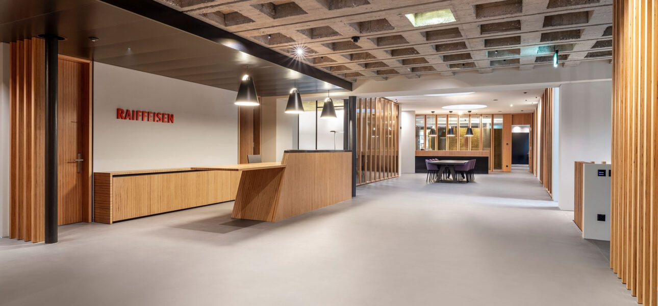 Industrial lobby interior with wooden reception desk, Raiffeisen sign, and seating area under a grid concrete ceiling with pendant lights.