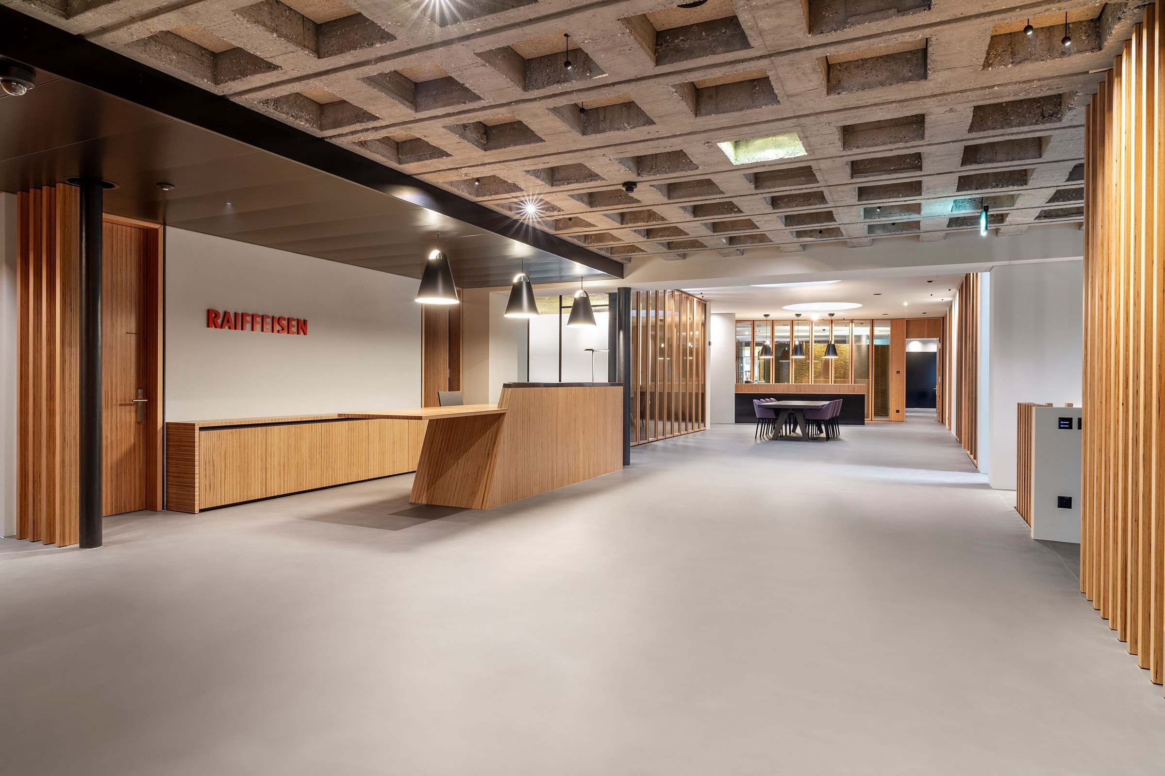 Industrial lobby interior with wooden reception desk, Raiffeisen sign, and seating area under a grid concrete ceiling with pendant lights.