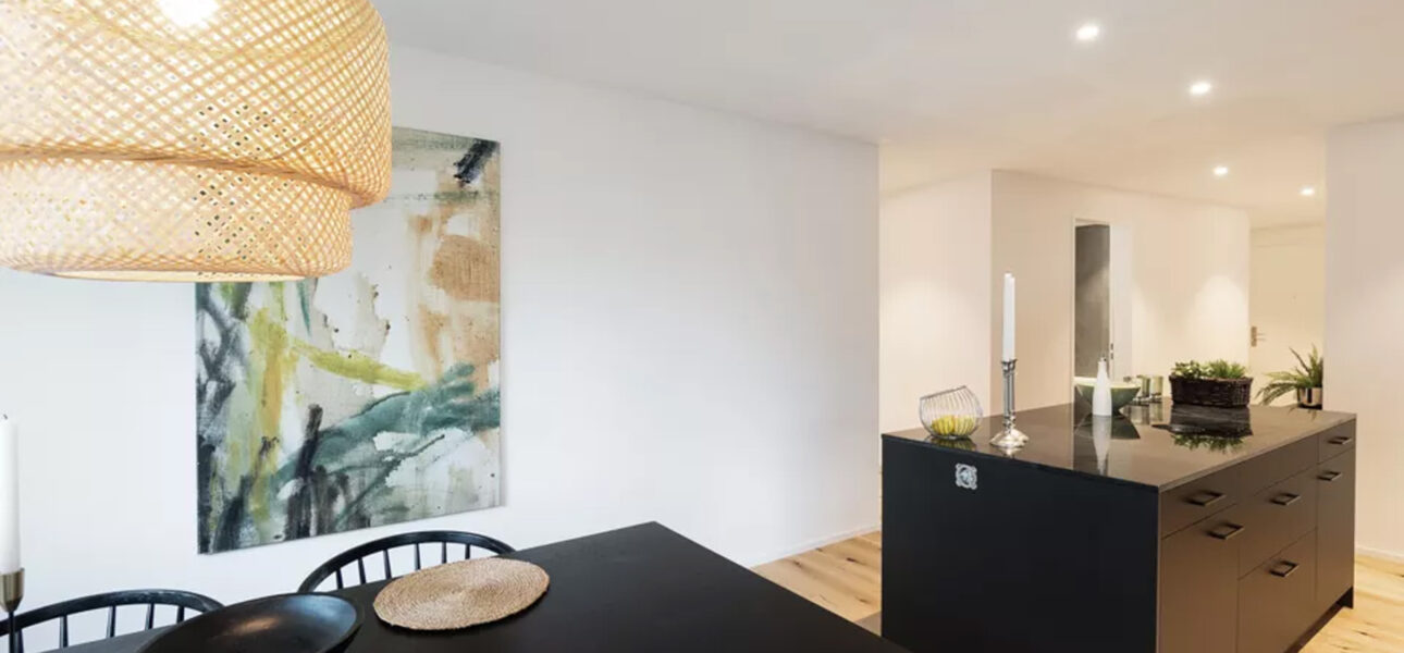 Open-concept dining area with a black table, three woven placemats, and a large woven pendant light overhead; abstract art on a white wall behind.